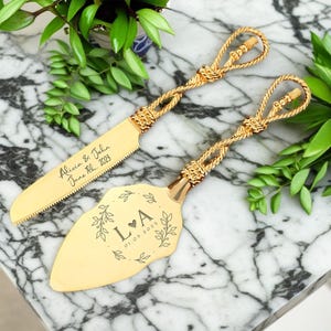 以下が含まれることがあります： 結び目の付いたハンドルが付いた金のウェディングケーキサーバーセット。サーバーには、「LA」のイニシャルと「01.03.2023」の日付が、花柄で囲まれた状態で刻印されています。ナイフには、「Alicia & John June 11th, 2023」と刻印されています。