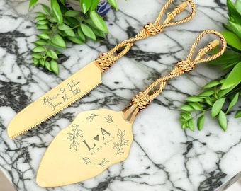May include: Gold wedding cake server set with a knotted handle. The server is engraved with the initials "LA" and the date "01.03.2023" surrounded by a floral design. The knife is engraved with "Alicia & John June 11th, 2023".
