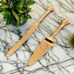 以下が含まれることがあります： 金色のケーキナイフとサーバーのセット。ナイフとサーバーは、花の模様の装飾的なハンドルが付いています。ナイフには「Hena & Rumel」という名前と「Dec 22nd, 2024」の日付が刻まれています。サーバーにはイニシャル「H R」と日付が刻まれています。