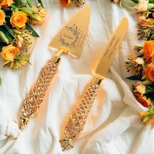 以下が含まれることがあります： 金色のケーキサーバーとナイフのセット。装飾的な格子模様のハンドルが付いています。サーバーには花の模様とイニシャルがあり、ナイフには名前と日付が刻まれています。白い布の上にオレンジ色の花と一緒に置かれています。