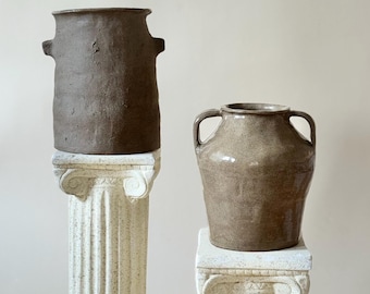 May include: Two ceramic vases in shades of brown and tan sit atop decorative white columns. The taller vase has two handles and a textured surface. The shorter vase has a smooth, glossy finish and two handles. The vases are displayed against a neutral background.