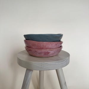 May include: A stack of three ceramic bowls in shades of blue and pink, resting on a small, light-colored wooden stool. The bowls have a textured surface and a simple, modern design. The stool has three legs.