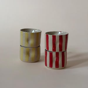 Puede incluir: Cuatro tazas de cerámica dispuestas en dos pilas. Una pila presenta rayas verticales amarillas sobre una base de color blanco roto moteado, mientras que la otra pila muestra rayas verticales rojas sobre una superficie de cerámica similar.