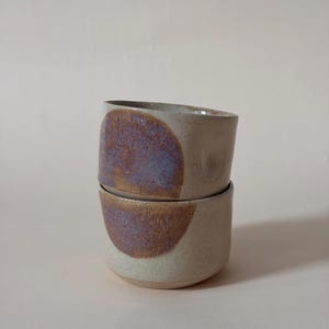 May include: Two stacked ceramic cups with a speckled off-white base and a prominent circular patch of textured, iridescent purple and brown glaze on the front of each vessel. The cups have a simple, cylindrical shape with a smooth, rounded rim.