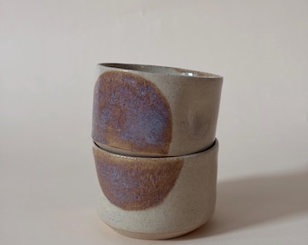 May include: Two stacked ceramic cups with a speckled off-white base and a prominent circular patch of textured, iridescent purple and brown glaze on the front of each vessel. The cups have a simple, cylindrical shape with a smooth, rounded rim.
