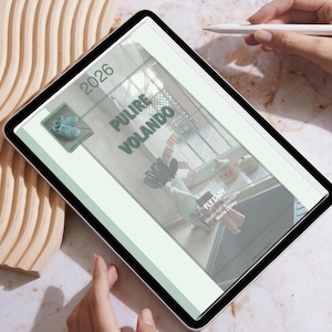 Może przedstawiać: Cyfrowy tablet wyświetlający stronę z tekstem "2026 PULIRE VOLANDO". Strona ma jasny zielony design z ilustracjami butów i przedmiotów kuchennych. Pióro jest trzymane w pobliżu ekranu.
