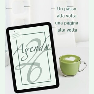 Può includere: Un tablet digitale che mostra la parola "Agenda" in un carattere elegante, con un bordo verde. Una tazza di matcha latte verde con un disegno a forma di cuore è accanto al tablet. Il testo "Un passo alla volta una pagina alla volta" è visibile.
