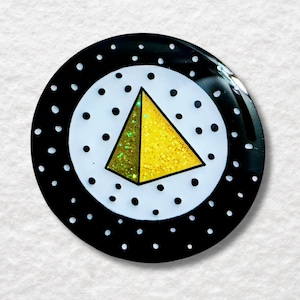 Pode incluir: Um broche circular com um centro branco de bolinhas pretas e um triângulo central de glitter amarelo. A borda externa é preta com bolinhas brancas. O design tem um acabamento brilhante e uma borda lisa e arredondada em toda a peça.
