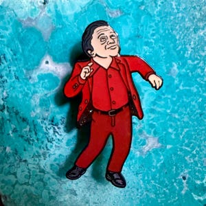 Könnte beinhalten: Ein Metall-Emaille-Pin mit einer Cartoon-Illustration einer Person mit dunklem Haar, die einen leuchtend roten Anzug, ein rotes Hemd und schwarze Schuhe trägt. Die Figur ist in einer dynamischen Pose mit erhobenem Arm dargestellt.