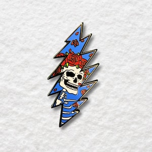 Puede incluir: Pin de rayo azul con una calavera adornada con rosas rojas. El rayo tiene un contorno dorado y un fondo azul con un patrón floral. La calavera tiene una cara blanca y detalles negros.