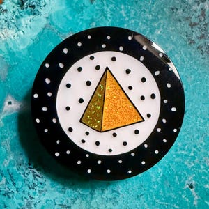 Peut inclure: Une broche circulaire présentant un disque central blanc à pois noirs et une pyramide à paillettes dorées. La bordure extérieure est noire avec des pois blancs. Le design est plat et brillant, mettant en valeur une forme pyramidale géométrique.