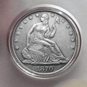 May include: A silver coin featuring a seated woman holding a shield with the word "LIBERTY" and a staff. The coin is dated 1870 and surrounded by stars and a beaded border. The coin is in a circular frame.
