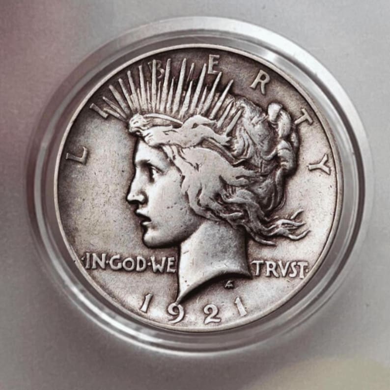 May include: A silver coin from 1921, featuring a profile of a woman with a radiant crown. The coin is encased in a clear protective holder. Inscriptions include "LIBERTY", "IN GOD WE TRUST", and "1921".