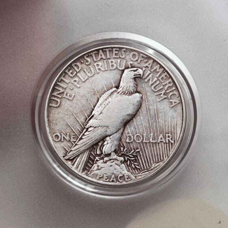May include: A silver one-dollar coin with an eagle design, set in a silver bezel. The coin features the words "UNITED STATES OF AMERICA", "E PLURIBUS UNUM", "ONE DOLLAR", and "PEACE". The eagle is perched on a rock.