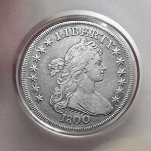Może przedstawiać: Srebrna moneta z napisem "LIBERTY" u góry i rokiem "1800" u dołu. Moneta przedstawia profil kobiety otoczony gwiazdami i ozdobnym obramowaniem.