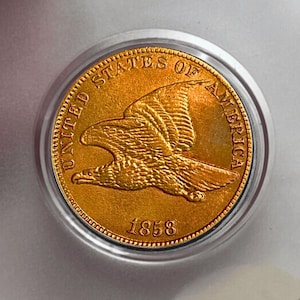 May include: A gold-coloured coin depicting a flying eagle. The coin is inscribed with "UNITED STATES OF AMERICA" and the year "1858". The coin is encased in a clear protective holder.