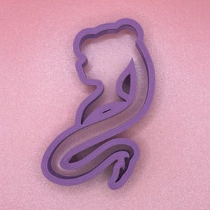 Puede incluir: Cortador de galletas de plástico morado con forma de sirena con una cola larga y fluida.