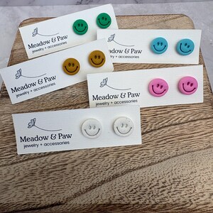 May include: A collection of colorful smiley face stud earrings on display. The earrings are in various colors, including green, yellow, blue, pink, and white. Each pair is mounted on a small white card with the text "Meadow & Paw jewelry + accessories".