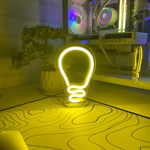 May include: A yellow neon light shaped like a light bulb on a black base. The light is on a yellow and black patterned surface.