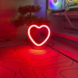 Op de afbeelding: Een rood neon hartvormig licht met een witte omtrek, dat op een wit oppervlak met een rood en zwart patroon staat.