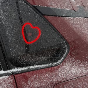 May include: A red car with a heart drawn on the back window. The car is covered in a light dusting of snow.