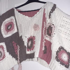 Könnte beinhalten: Ein handgefertigtes, offen geschnittenes Häkeltop im Patchwork-Design. Das Top hat Quadrate und Blumenmotive in Weiß, Braun und Rosa. Der Ausschnitt und die Kanten haben eine gewellte Bordüre. Die Ärmel sind weit.