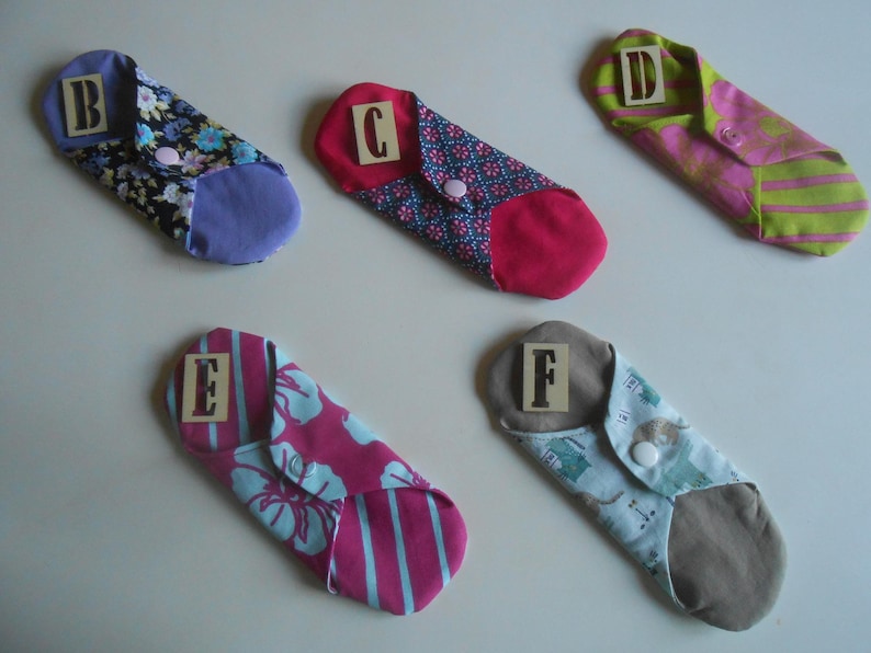 May include: Six cloth menstrual pads with different patterns and colors. Each pad has a letter label, from A to F, sewn on the front.