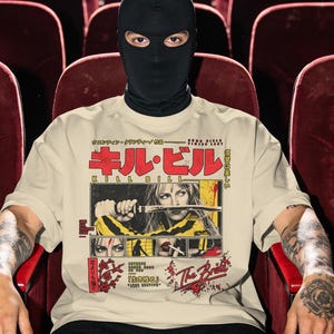 May include: A beige short-sleeved t-shirt featuring a graphic print from the film Kill Bill. The design includes Japanese text, the title Kill Bill, and multiple panels showing a character holding a sword, a silhouette, and red floral accents.