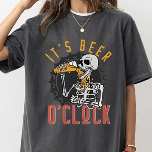 May include: A dark gray t-shirt with a graphic of a skeleton drinking beer. The text on the shirt reads "It's Beer O'Clock".