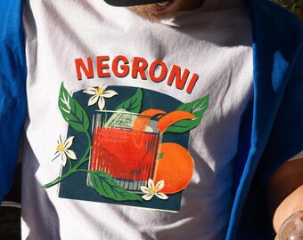 May include: White t-shirt with a graphic of a Negroni cocktail, featuring an orange slice, ice cubes, and a sprig of orange blossoms.