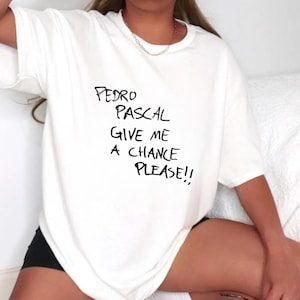 Op de afbeelding: Wit oversized T-shirt met zwarte tekst die "Pedro Pascal Give Me A Chance Please!!" zegt.