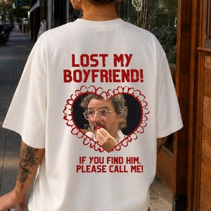 Peut inclure: Un t-shirt blanc avec du texte rouge indiquant LOST MY BOYFRIEND! IF YOU FIND HIM, PLEASE CALL ME! Le motif comprend un cadre en forme de cœur avec un bord festonné contenant une image photographique d'une personne portant des lunettes.