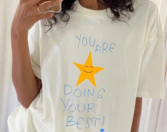 May include: A white t-shirt with a blue and yellow graphic design. The graphic features a smiling yellow star and the text "You are doing your best!"