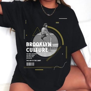 Può includere: T-shirt nero con le parole "BROOKLYN CULTURE" in bianco e giallo. La maglietta presenta un'immagine in bianco e nero di una donna in costume da bagno. Il testo aggiuntivo include "YOU ONLY LIVE ONCE", "404 NOT FOUND" e "THE SKY IS THE LIMIT".