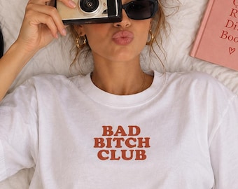 May include: A white short-sleeved t-shirt featuring the text BAD BITCH CLUB printed in a bold, rust-coloured font across the chest. The shirt has a classic crew neckline and a relaxed fit, providing a simple and clean aesthetic for casual wear.