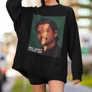 May include: Black sweatshirt with a portrait of a man and the text "BUT DADDY I LOVE HIM". The sweatshirt is paired with black shorts, white socks, and trainers. A black beanie is worn on the head.