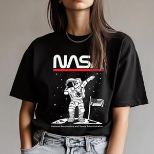 May include: Black t-shirt with a white NASA logo and a white illustration of an astronaut doing the dab on the moon. The text "National Aeronautics and Space Administration" is printed below the image.