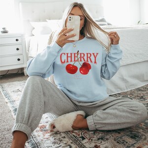 May include: A light blue sweatshirt with the word "CHERRY" printed in red letters. Two red cherries are printed below the word. The cherries have stems and leaves.
