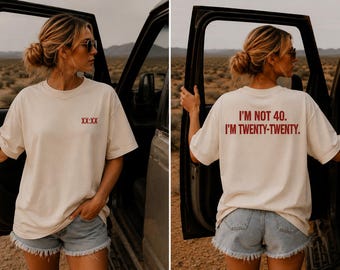 May include: A cream-colored short-sleeve t-shirt featuring the red text XX:XX on the front chest and the red text I'M NOT 40. I'M TWENTY-TWENTY. printed on the back. The shirt has a crew neckline and a relaxed fit, paired with light blue denim shorts.