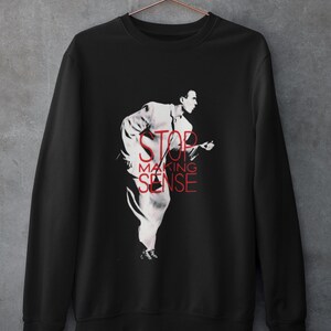 May include: A black long-sleeve sweatshirt featuring a white graphic of a person in a suit running, overlaid with the red text STOP MAKING SENSE. The garment is displayed on a copper-colored wire hanger against a gray textured wall.