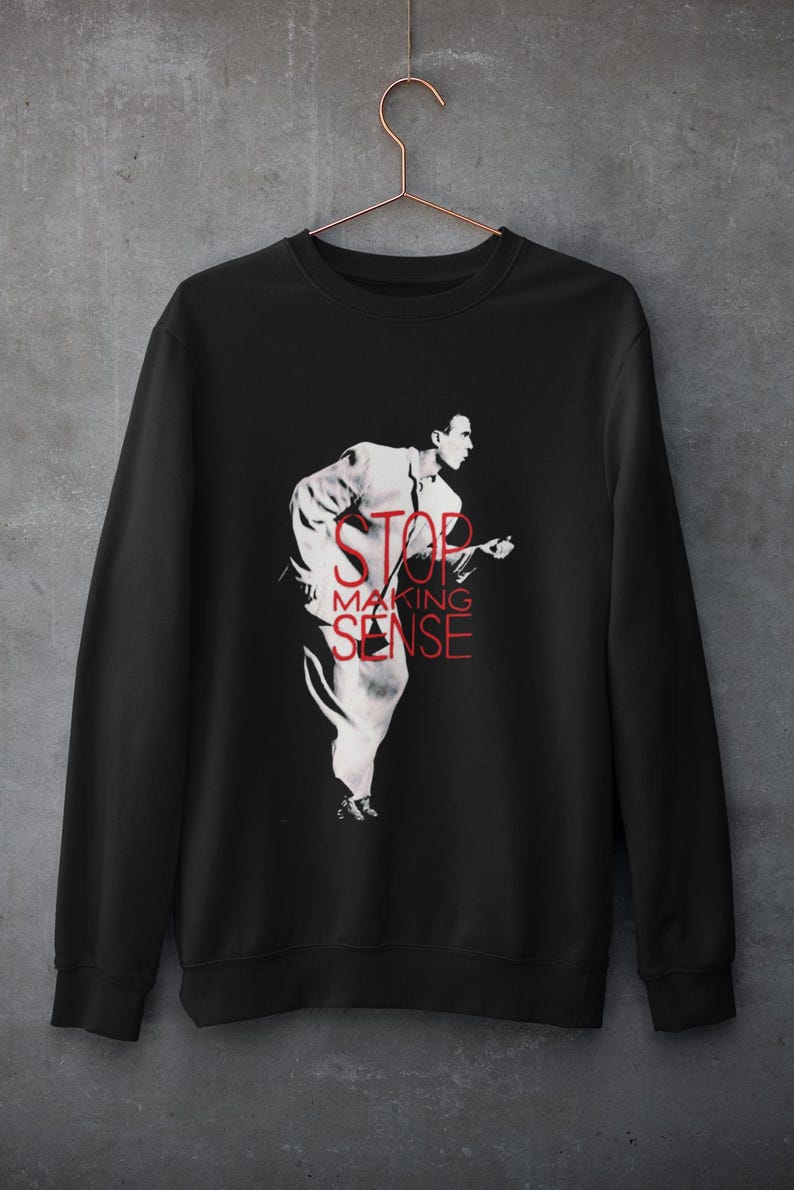 May include: A black long-sleeve sweatshirt featuring a white graphic of a person in a suit running, overlaid with the red text STOP MAKING SENSE. The garment is displayed on a copper-colored wire hanger against a gray textured wall.