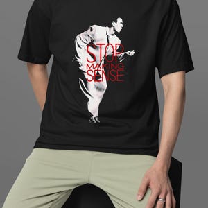 May include: Black t-shirt featuring a red and white graphic of a person playing a guitar, with the text "STOP MAKING SENSE". The person is wearing a white suit. The shirt is being worn by a person wearing light green pants.