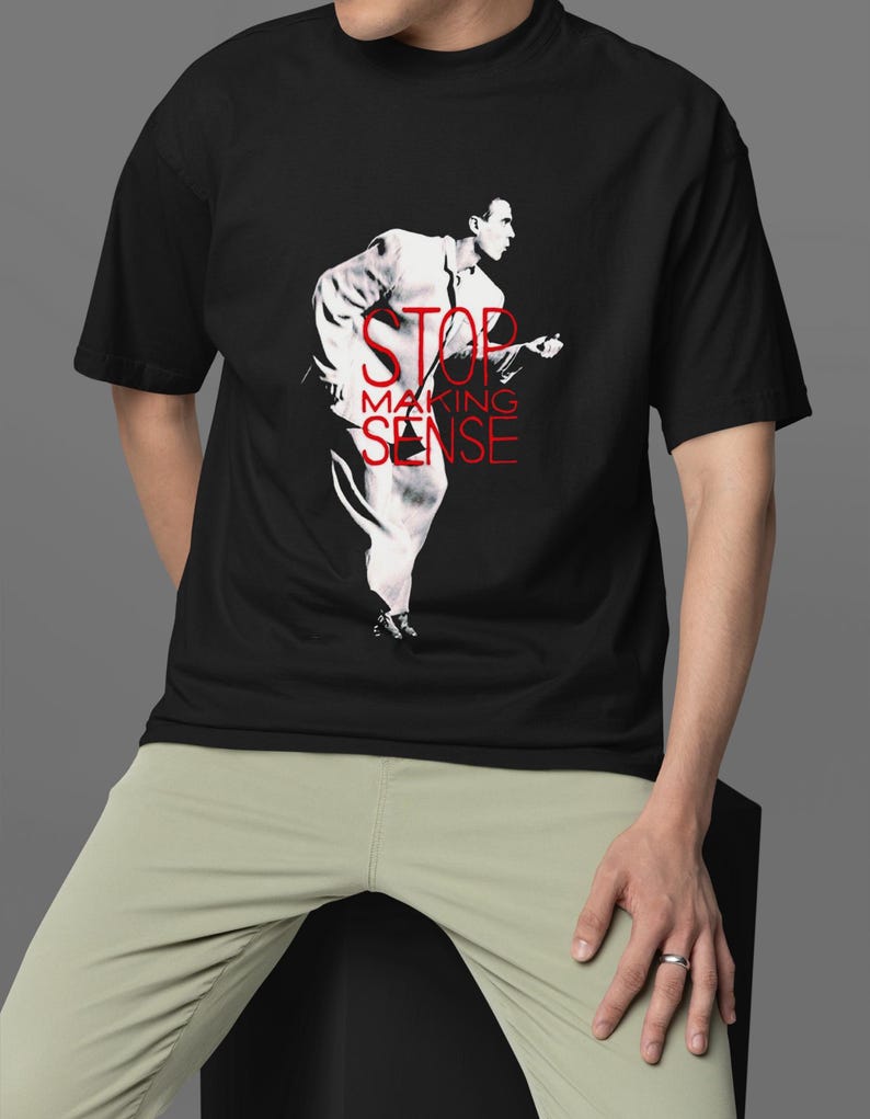 May include: Black t-shirt featuring a red and white graphic of a person playing a guitar, with the text "STOP MAKING SENSE". The person is wearing a white suit. The shirt is being worn by a person wearing light green pants.