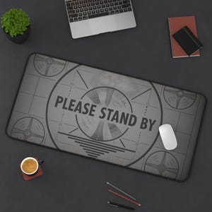 以下が含まれることがあります： レトロなテレビのテストパターンデザインと「PLEASE STAND BY」の文字が入った灰色の長方形のデスクマット。ラップトップ、マウス、コーヒーカップも机の上にあります。小さな植木鉢とノートも表示されています。
