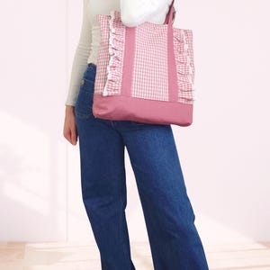 May include: A pink and white gingham tote bag featuring vertical pink fabric straps, ruffled side panels with white lace trim, and a solid pink base. The bag has two shoulder straps and is constructed from a combination of patterned and solid fabric.
