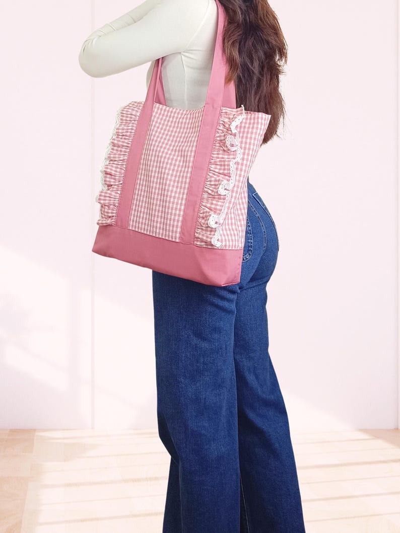 May include: A pink and white gingham tote bag featuring a solid pink base and two matching pink shoulder straps. The bag is accented with vertical white lace trim ruffles along the sides of the gingham panels, creating a textured appearance.