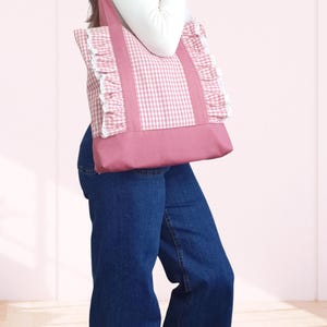 May include: A tote bag featuring a pink and white gingham pattern on the upper section and a solid pink base. The bag includes two shoulder straps and vertical ruffles along the sides, accented with white lace trim along the ruffle edges.