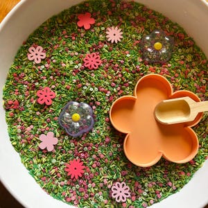 May include: A sensory bin filled with green and pink sprinkles, small pink flower-shaped cutouts, and two clear flower-shaped toys with yellow centers. An orange flower-shaped mold and a small wooden scoop are also present.