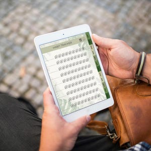 May include: A white tablet displaying a habit tracker app. The app features a green and white leaf design and a grid of check boxes. The tablet is held by a person, with a brown leather bag in the background.