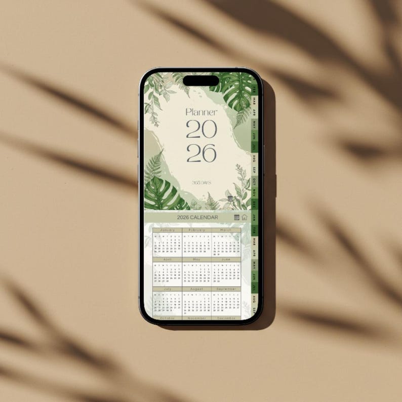 May include: A smartphone displays a digital planner for the year 2026. The screen features a calendar layout with a botanical design, including green leaves and the text "Planner 2026". The phone is set against a neutral background.